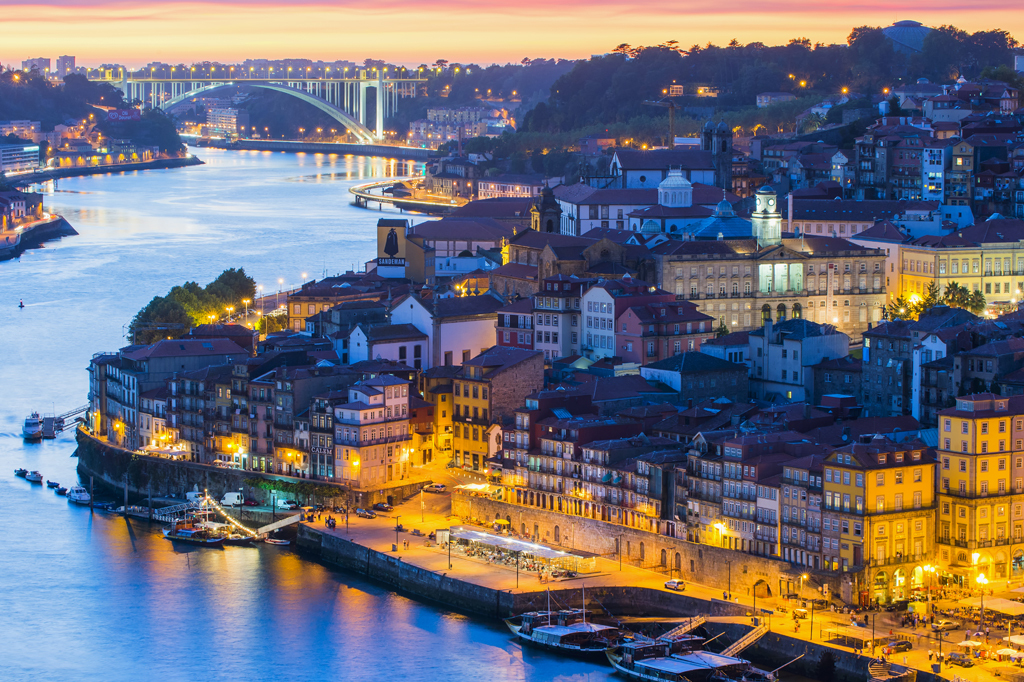 The city of Porto by evening, bathed in warm lights next to the river Douro. A majestic bridge sits across the horizon.